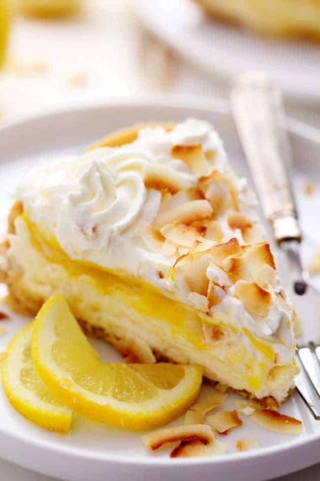 No Bake Lemon Macaroon Cheesecake is a coconut filled cheesecake on top of a golden oreo crust. Topped with lemon curd andย fresh whipping cream this easy to make dessert will be a huge hit! A slice of No Bake Lemon Macaroon Cheesecake on a white plate with a metal spoon and some fresh cut lemon on the side.