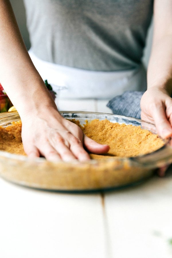 This Easy Lemon Pudding Pie is a creamy & light, slightly tangy, and very refreshing 3 layer dessert!ย Its buttery graham cracker crust is topped with a sweet and tart filling of lemon pudding and whipped cream.ย Insanely delicious and so quick and easy to make! A hand pressing down the pie crust in the clear pie pan.