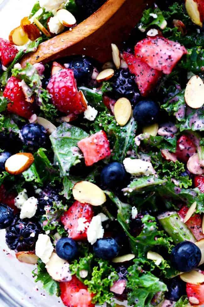 Triple Berry Kale Salad with Creamy Strawberry Poppyseed Dressing is made with delicious fresh summer berries and is tossed with kale, crunchy almonds and topped with feta cheese. A healthy and light salad that is perfect for summer! Close up photo of Triple Berry Kale Salad with Creamy Strawberry Poppyseed Dressing in a clear bowl with a wooden spoon.