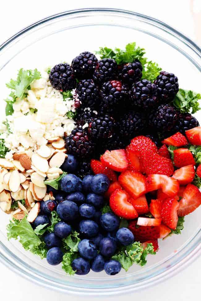Triple Berry Kale Salad with Creamy Strawberry Poppyseed Dressing is made with delicious fresh summer berries and is tossed with kale, crunchy almonds and topped with feta cheese. A healthy and light salad that is perfect for summer! Triple Berry Kale Salad ingredients in a large clear bowl.