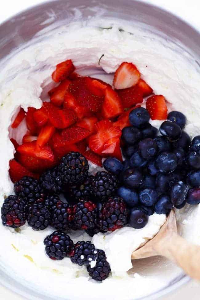 No Bake Very Berry Cheesecake comes together in just 10 minutes! It is filled with fresh summer berries hidden inside a creamy cheesecake and is incredible! Strawberries, blackberries, and blueberries being mixed into the cheesecake mixture in a large mixing bowl.