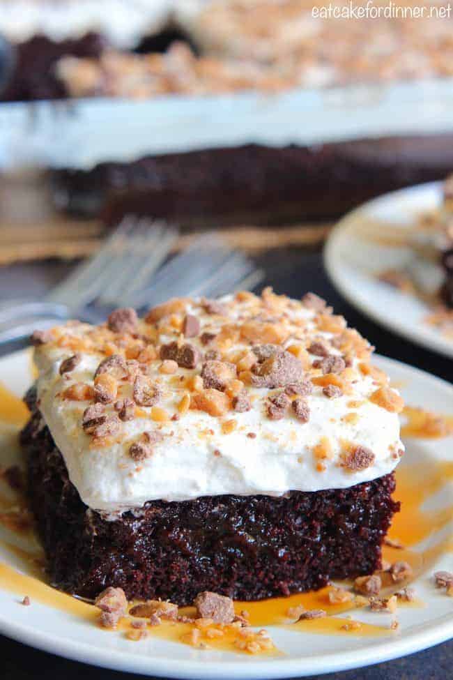Better than Anything Zucchini Poke Cake has caramel inside of a moist chocolate cake. Topped with cool whip and rich toffee bits this will be a HUGE hit! You will never know zucchini is inside!ย Better than Anything Zucchini Cake on a white plate.