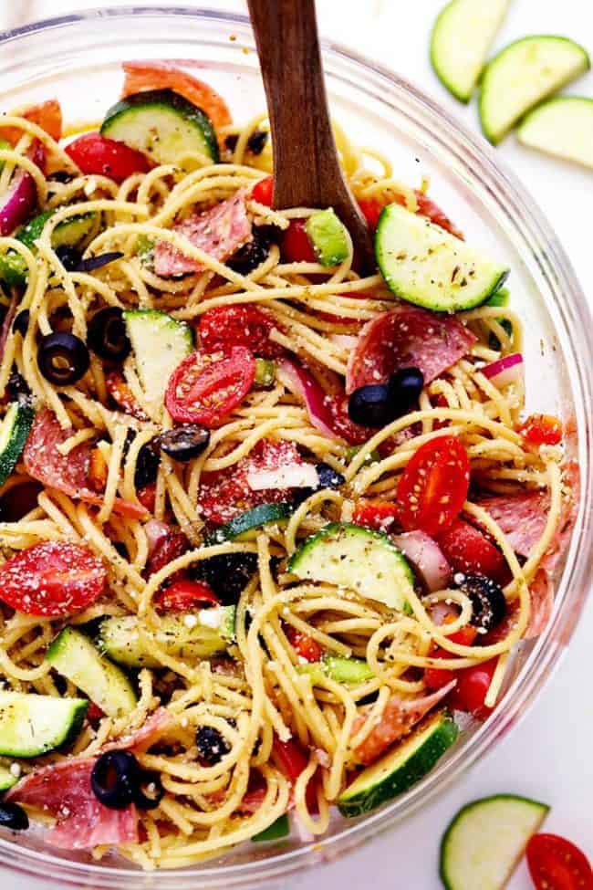 Italian Spaghetti Salad is made with spaghetti, fresh summer veggies, salami and tossed in a homemade zesty Italian dressing. This is a summer potluck must! Italian Spaghetti Salad in a clear bowl with a wooden spoon.