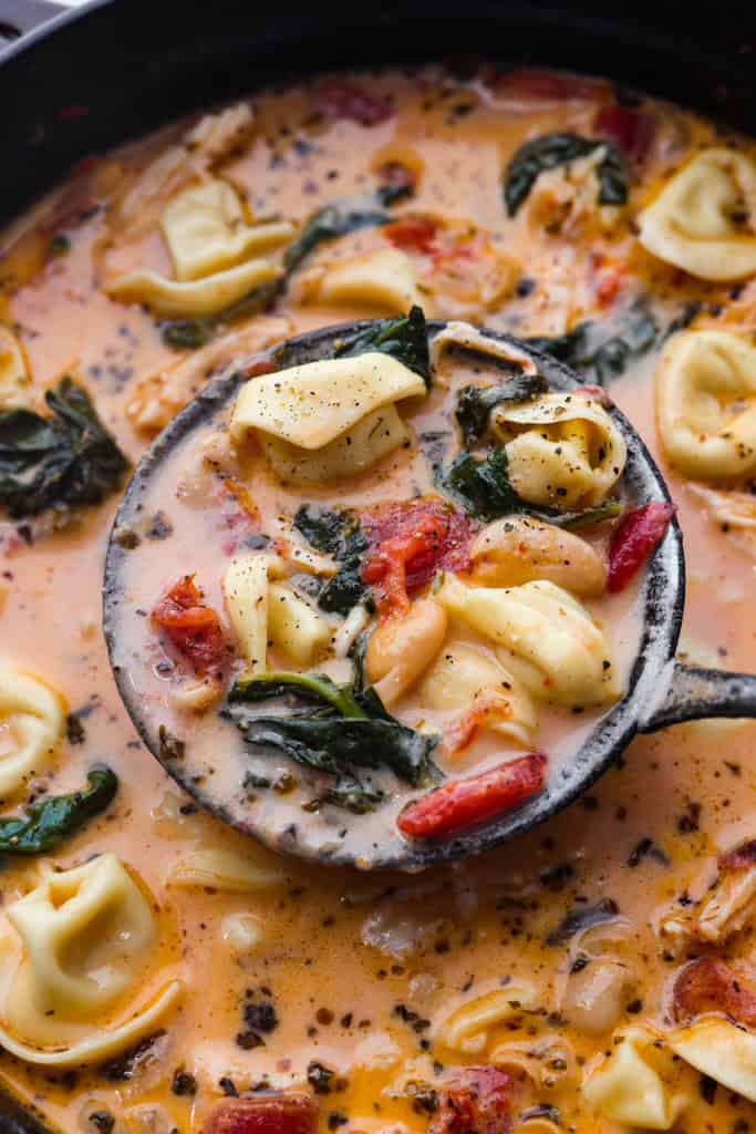 Creamy Tuscan garlic tortellini soup is so easy to make and one of the best soups that you will make! This soup is loaded with cheesy tortellini, diced tomatoes healthy spinach and hearty white beans. This soup is so creamy and delicious, I know your family will love it! Close up shot of a ladle scooping a scoop of creamy Tuscan garlic tortellini soup above the pot.