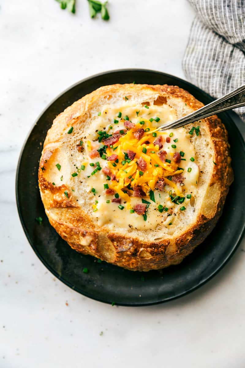 Creamy and cheesy red potato soup that the whole family will love! Red Potato Soup in a bread bowl with a spoon in the soup. All sitting on a black plate.