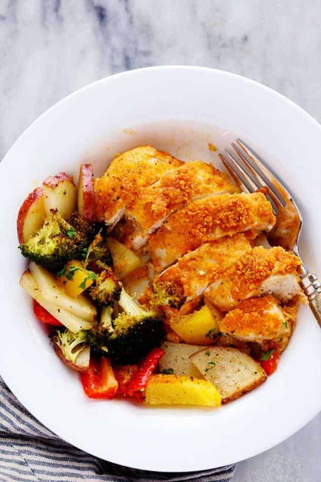 One Pan Crispy Parmesan Paprika Chicken with Vegetables has tender and juicy chicken with a crispy parmesan paprika breading surrounded by veggies. It it is full of flavor and makes an amazing one pan dinner your family will love! Crispy Parmesan paprika chicken cut into slices and type of vegetables and a white bowl with a fork on the side.