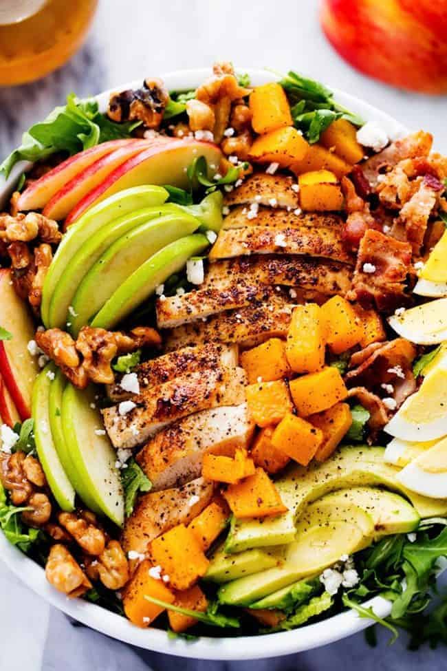 Harvest Cobb Chicken Salad with a Honey Apple Cider Vinaigrette is filled with crisp fall apples, roasted butternut squash, and all of the delicious things of fall come together in this mouthwatering salad. Harvest cup chicken salad with a honey apple cider vinegar and a white bowl. On top of the salad or apple slices chicken, sliced avocados and hard boiled eggs.