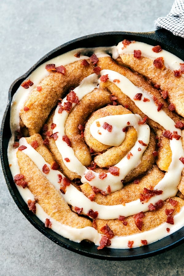 Skillet Maple Bacon Cinnamon Roll is an easy to make cinnamon roll skillet cake with maple cream cheese frosting and crumbled bacon. The perfect holiday breakfast! Maple bacon cinnamon roll in a black skillet with frosting overtop and garnished with bacon bits.