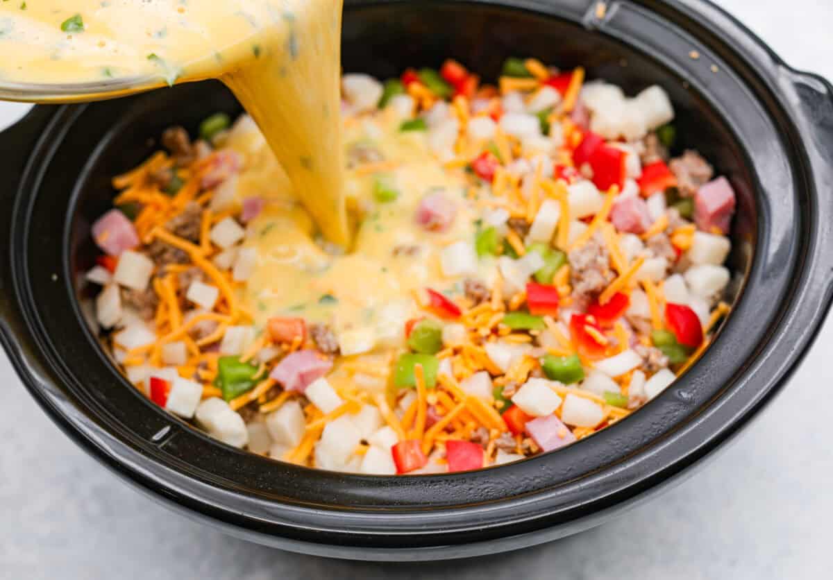 This Slow Cooker Breakfast Casserole is quick and easy to put together! Breakfast can be cooked in the slow cooker while you're sleeping! This casserole is loaded up with seasoned sausage, diced hash browns, tender vegetables, eggs, and cheese. Angel shot of someone pouring the egg mixture in to the veggie, meat and cheese mixture in the slow cooker.