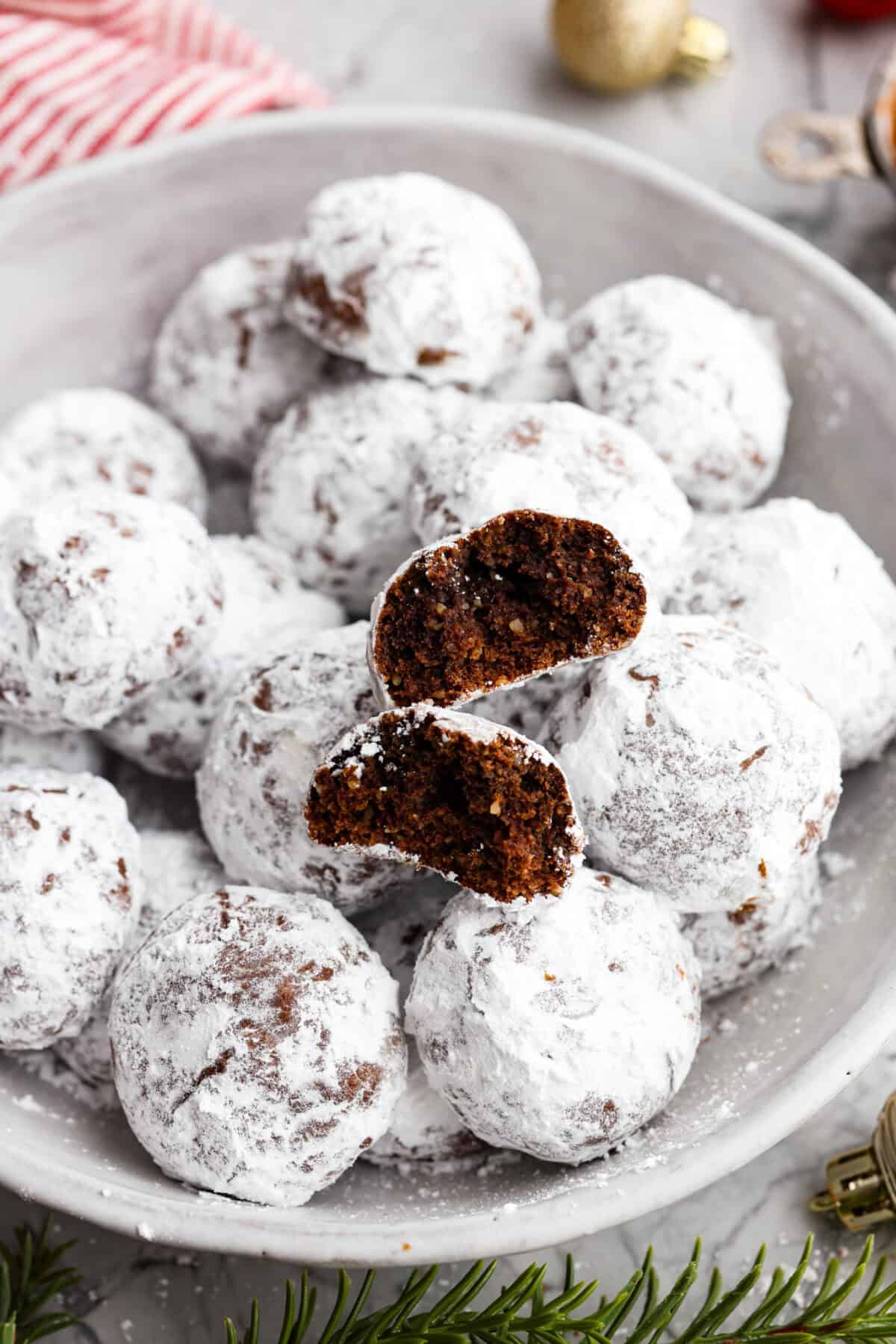 Chocolate Snowball Cookies are tender chocolate cookies with the best texture! A buttery cookie made with cocoa powder and ground walnuts gets coated in sweet powdered sugar. Make this classic holiday cookie a tradition every year! Close view of chocolate snowball cookies in a serving bowl with a cookie broken in half.