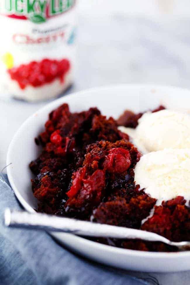Slow Cooker Cherry Chocolate Hot Fudge Cake is a perfectly tender chocolate cake with hidden cherries inside. It creates a magical fudge sauce while it is in the slow cooker and is completely irresistible! Slow cooker cherry chocolate half page cake in a white bowl with two scoops of ice cream on top and a spoon on the side.