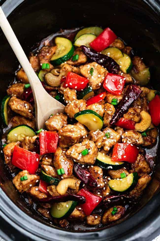 Skinny Slow Cooker Kung Pao Chickenย is aย delicious chicken coated in a sweet and spicy sauce with tender vegetables and crunchyย cashews. Skip the takeout, this is so muchย healthier and better! Skinny Slow Cooker Kung Pao Chicken in a slow cooker with a wooden spoon.