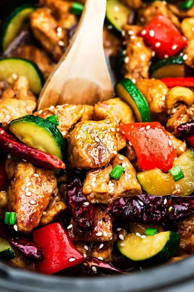 Skinny Slow Cooker Kung Pao Chickenย is aย delicious chicken coated in a sweet and spicy sauce with tender vegetables and crunchyย cashews. Skip the takeout, this is so muchย healthier and better! Skinny Slow Cooker Kung Pao Chicken unclose photo with a wooden spoon.