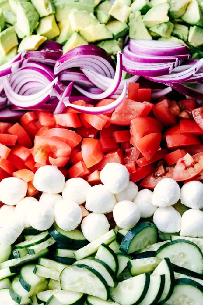 Italian Avocado Cucumber Tomato Salad is a fresh and delicious salad filled with avocado, tomato, red onion, cucumber and mozzarella cheese balls. Tossed in a tangy lemon Italian glaze and topped with feta cheese, you won’t be able to get enough! Italian Avocado Cucumber Tomato Salad ingredients in rows.