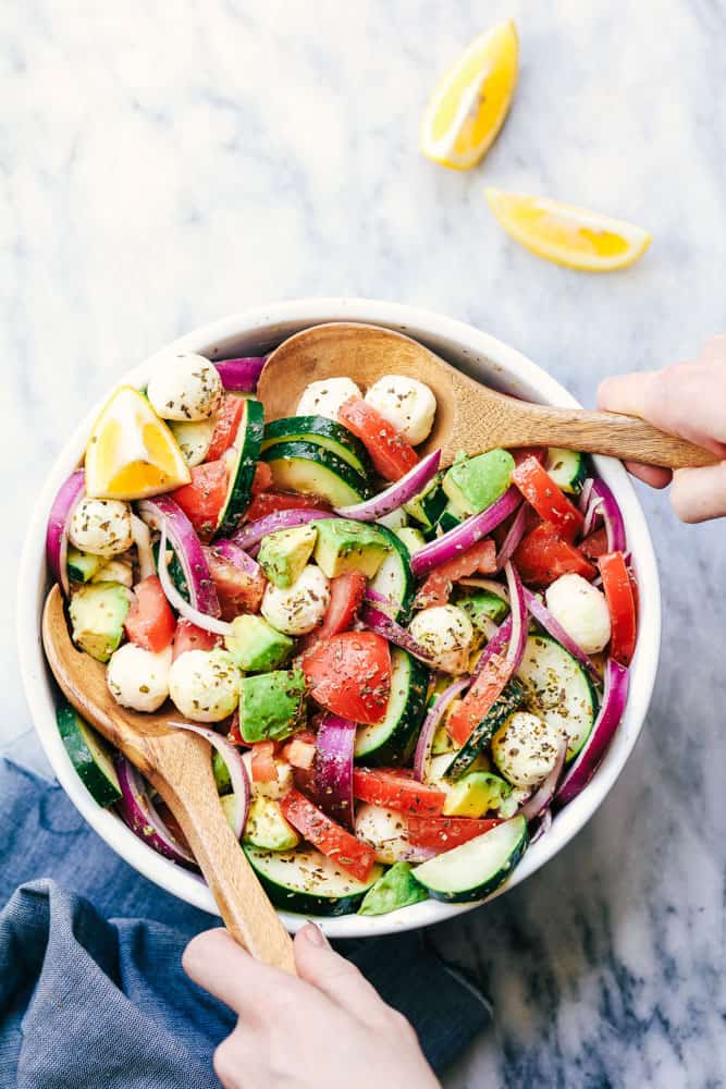 Italian Avocado Cucumber Tomato Salad is a fresh and delicious salad filled with avocado, tomato, red onion, cucumber and mozzarella cheese balls. Tossed in a tangy lemon Italian glaze and topped with feta cheese, you won’t be able to get enough! Italian Avocado Cucumber Tomato Salad in a white bowl with 2 wooden serving spoons.