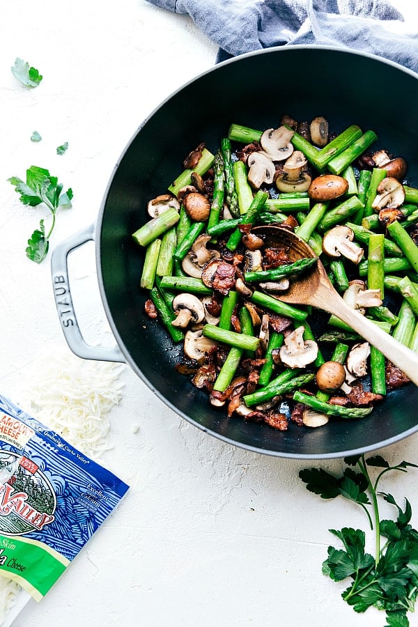 Cheesy Chicken, Asparagus, Bacon, and Mushroom Shells are made in one pan with an incredibly creamy white cheese sauce. This is a quick and easy meal that will become a new family favorite!ย Cheesy Chicken, Asparagus, Bacon, and Mushroom Shells in a skillet being stirred with a wooden spoon.