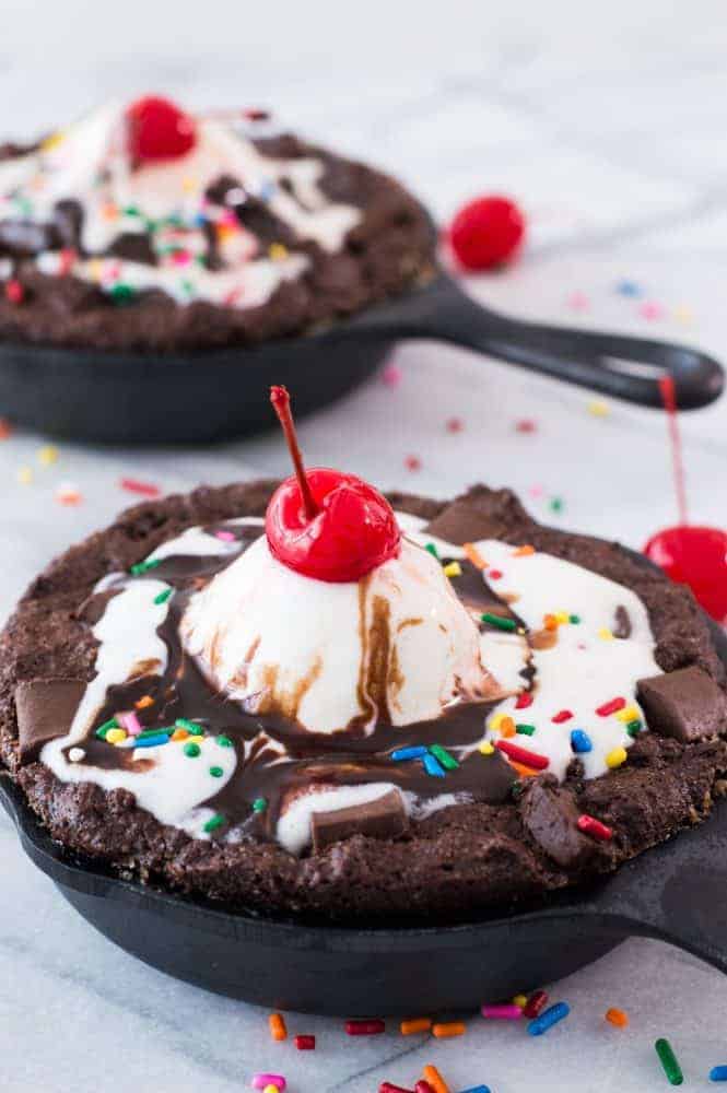 Skillet Brownie Sundaes! ย These mini skillets are filled with warm fudgy brownies loaded with chunks of chocolate and topped with ice cream, chocolate syrup, sprinkles and of course the cherry on top! Perfect small batch dessert for two!ย Skillet Brownie Sundaes in cast iron skillets close up.
