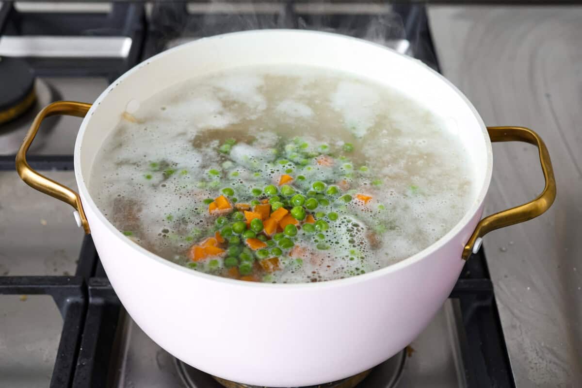 Creamy Chicken Noodle Casserole is comfort food at its finest! Soft and slightly chewy noodles, tender chicken, crisp vegetables, creamy and cheesy sauce, with a buttery cracker topping. This will become a family favorite! Angle shot of the noodles with peas and carrots in a pot with bowling water.