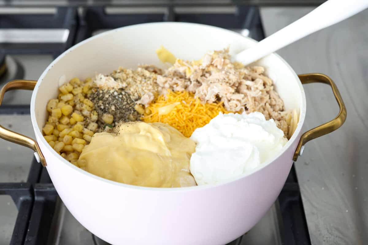 cooked chicken, corn, cream of chicken soup, milk, sour cream, onion flakes, Italian seasoning, garlic powder, salt, pepper, and cheddar cheese. Angle shot of the cooked chicken, corn, cream of chicken soup, milk, sour cream, onion flakes, Italian seasoning, garlic powder, salt, pepper, and cheddar cheese added to the pot with the drained noodles and veggies.