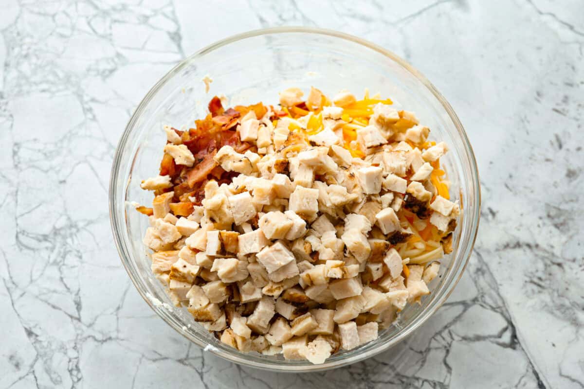 Chicken Bacon Ranch Pinwheels are an easy wrap your family or party guests will love! They are rolled up and loaded with hearty chicken, savory bacon, and cheese, all slathered in a delicious ranch-flavored cream cheese. You can enjoy them both hot and cold! Overhead shot of chicken, bacon and cheese over the cream cheese mixture in a bowl.