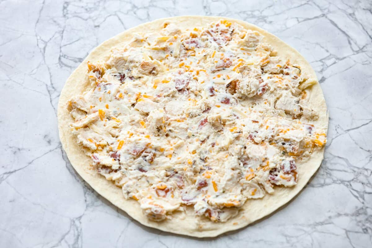 Chicken Bacon Ranch Pinwheels are an easy wrap your family or party guests will love! They are rolled up and loaded with hearty chicken, savory bacon, and cheese, all slathered in a delicious ranch-flavored cream cheese. You can enjoy them both hot and cold! Overhead shot of tortilla with filling mixture spread over the top.