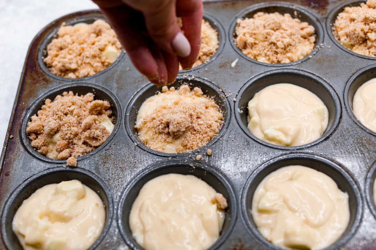 Apple Crumb Muffins are sweet and fluffy bakery-style muffins topped with a brown sugar crumble.ย Every bite is loaded with fresh apple pieces and spiced to perfection! Sixth photo of the crumb topping sprinkling on top of the muffin batter.