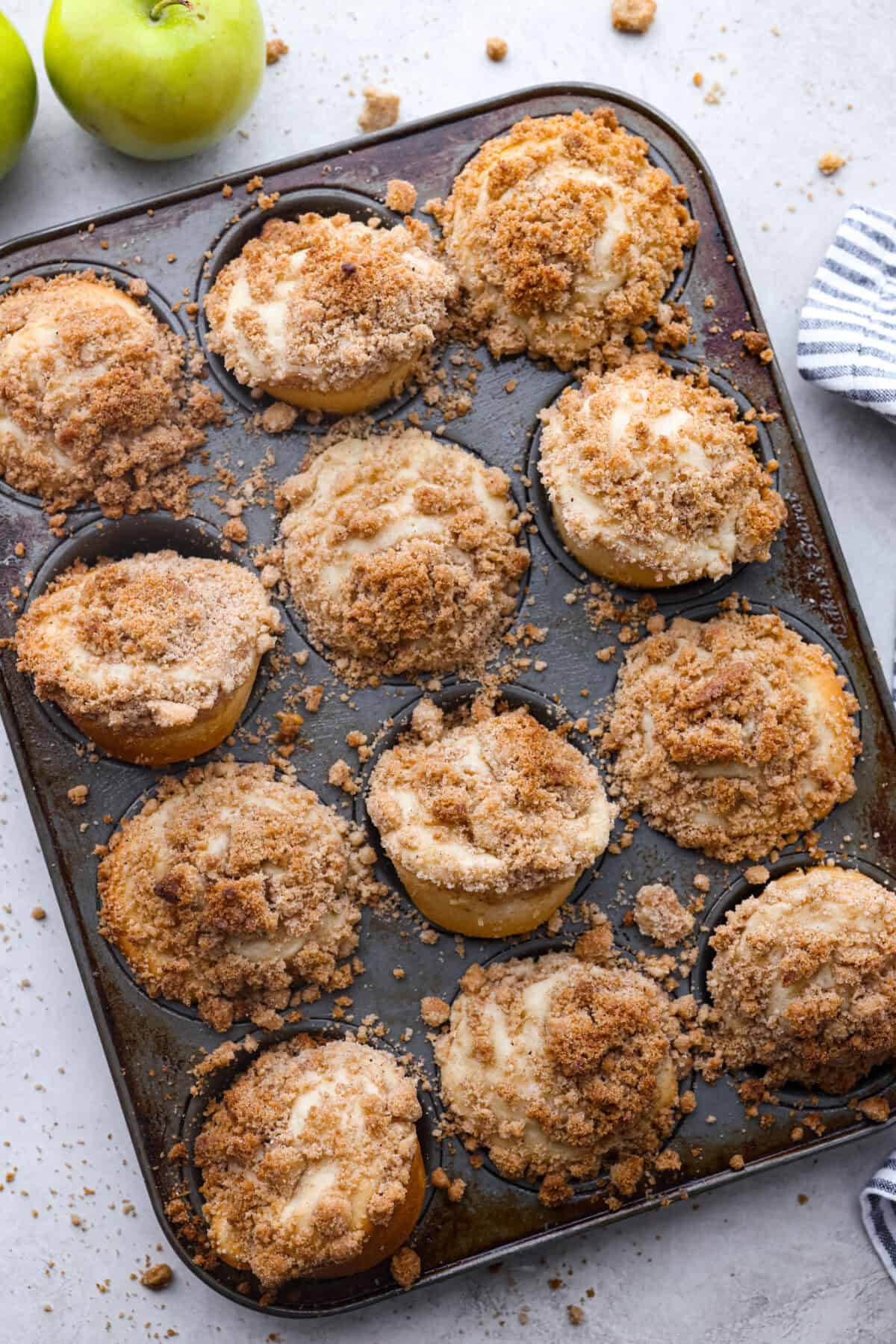 Apple Crumb Muffins are sweet and fluffy bakery-style muffins topped with a brown sugar crumble.ย Every bite is loaded with fresh apple pieces and spiced to perfection! Top view of apple crumb muffins in a muffin pan.