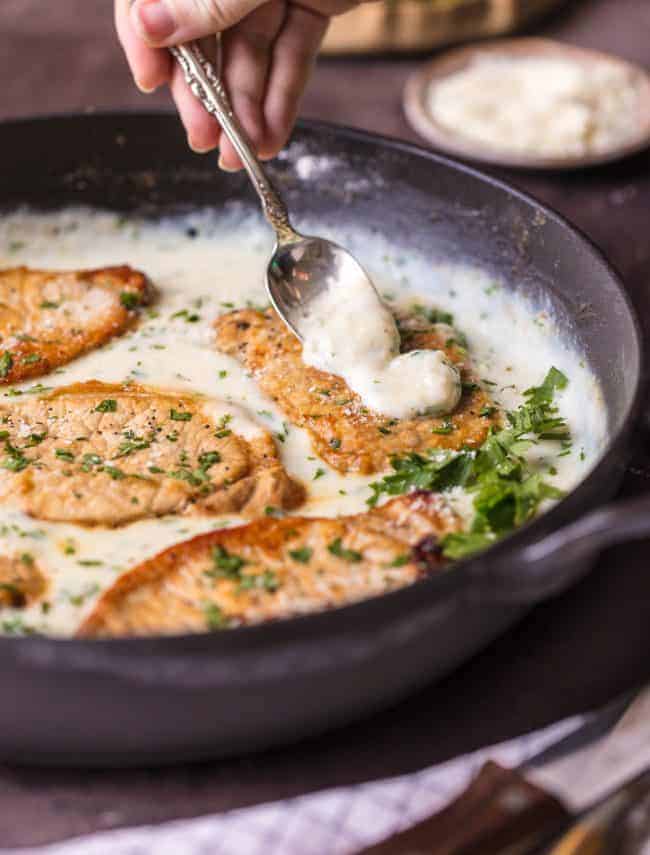 These Creamy Parmesan Pork Chops are sure to be a hit around the family table. The simple garlic parmesan sauce is oh so delicious and so easy anyone can make it in minutes.ย These Creamy Parmesan Pork Chops with a spoon pouring sauce over the pork chops in a skillet.