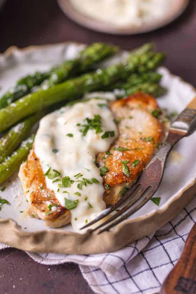 These Creamy Parmesan Pork Chops are sure to be a hit around the family table. The simple garlic parmesan sauce is oh so delicious and so easy anyone can make it in minutes.ย A plate of Creamy Parmesan Pork Chops with asparagus.