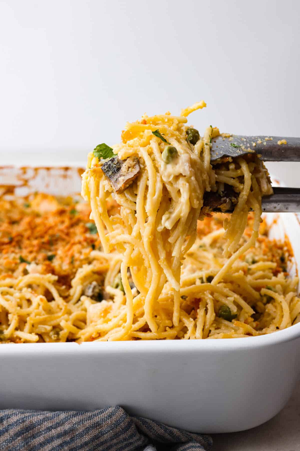 This chicken tetrazzini casserole is packed with delicious flavors and textures. It has hearty chicken, tender mushrooms, peas, and pasta, all tossed in a creamy, rich sauce. The casserole is topped with crispy, buttery breadcrumbs and a sprinkle of Parmesan cheese with the most irresistible crunch and flavor. Side shot of tongs lifting a portion of chicken tetrazzini casserole out of the casserole dish.