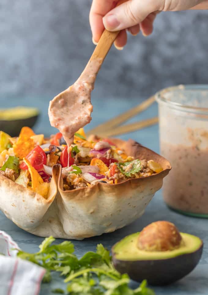 Thisย Creamy Taco Salad is the ultimate fun party meal for game day. It's simple, perfect for a crowd, and oh so delicious! Creamy Taco Salad with refried beans being drizzled over the top. There is a fresh cut avocado in the forefront and a jar of refried beans in the background.