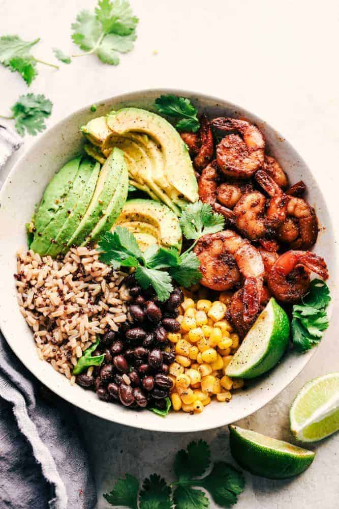 Blackened Shrimp Avocado Burrito Bowls are the perfect way to ring in the New Year with big bold flavor and a healthy meal! These are packed with blackened shrimp, brown rice, corn, black beans, and avocado. They are also great for meal prep! Blackened Shrimp Avocado Burrito Bowl in a white bowl with limes and cilantro on the side.