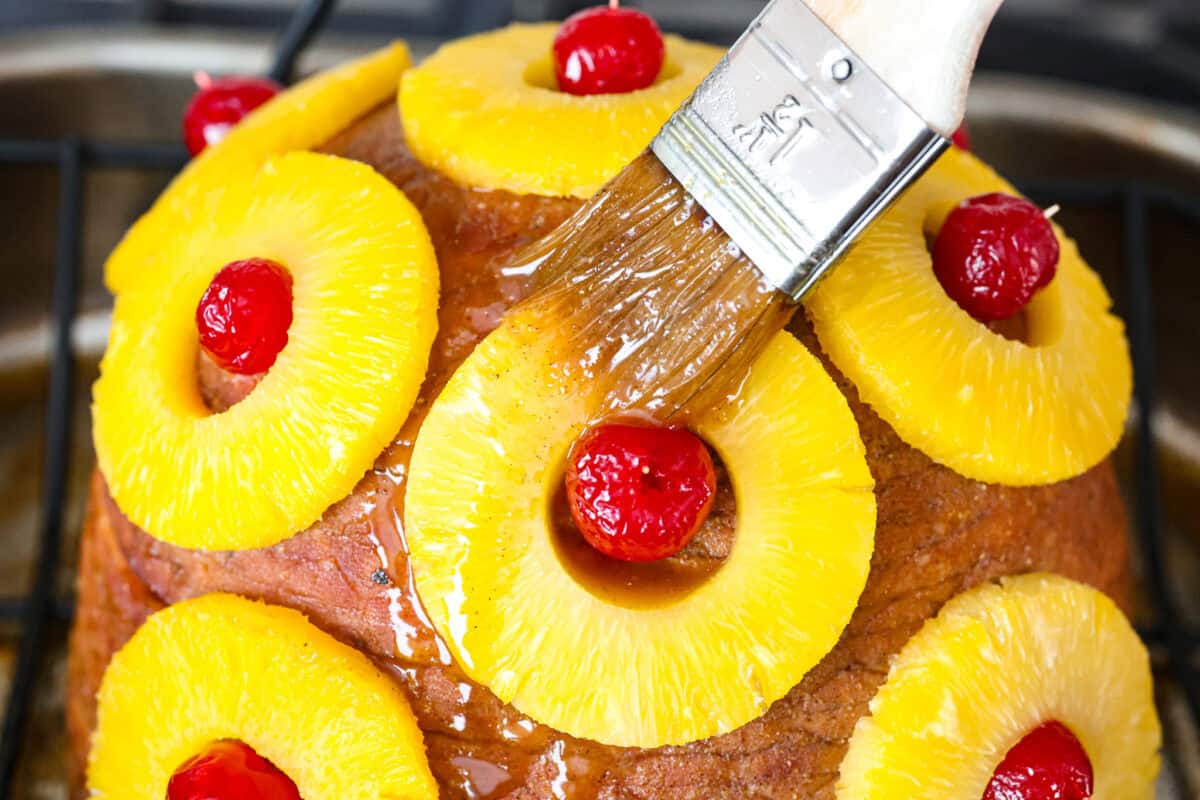 Brown Sugar Pineapple Glazed Ham is coated with a sweet and tangy spiced glaze that caramelizes on the edge of the ham. It's covered in juicy pineapple rings and maraschino cherries that add a tangy sweetness and juices to the succulent ham. Close up shot of someone putting a second coat of the pineapple glaze on the ham.