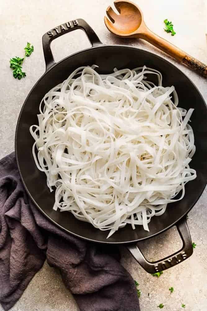 Mongolian Chicken Noodles - a one pot 30 minute meal perfect to curb those takeout cravings. Made with chicken, veggies, gluten free rice noodles and a delicious savory Asian-inspired sauce. Uncooked noodles in an iron skillet and a wooden spoon next to it.