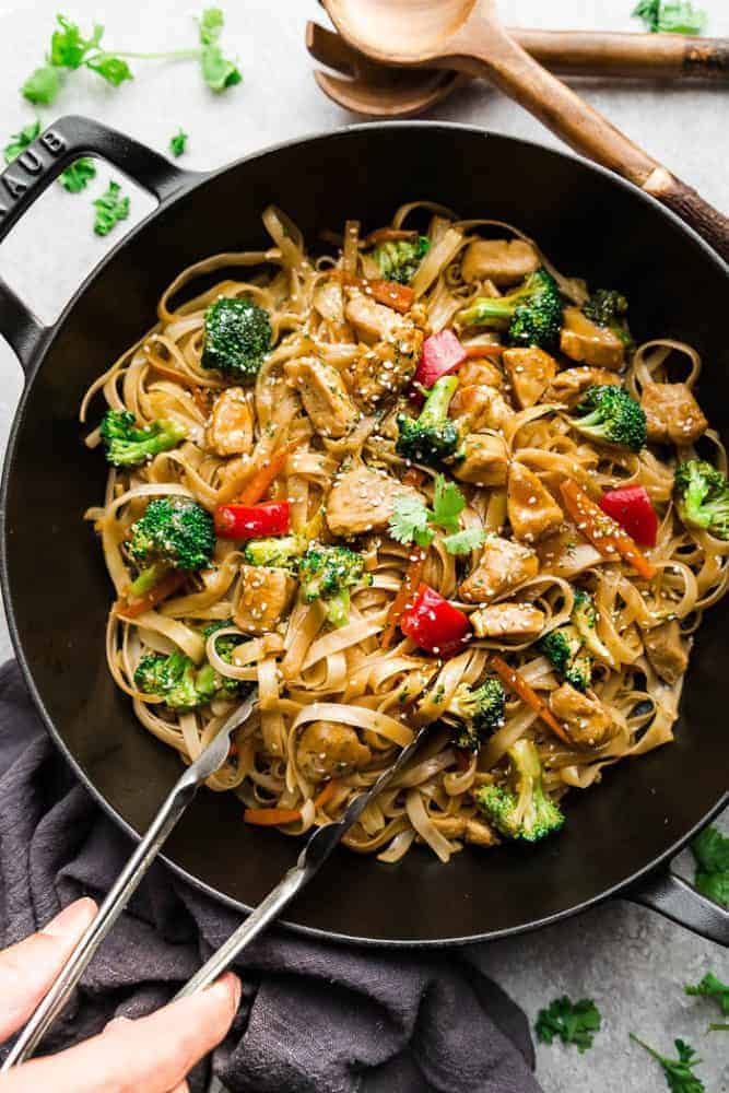 Mongolian Chicken Noodles - a one pot 30 minute meal perfect to curb those takeout cravings. Made with chicken, veggies, gluten free rice noodles and a delicious savory Asian-inspired sauce. Mongolian Chicken Noodles in a iron skillet with a set of tongs and wooden spoons.