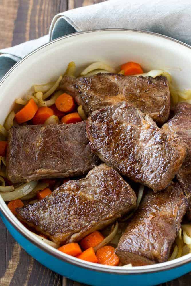 These fork tender root beer glazed short ribs are the perfect meal for a cold winter night. Serve them over mashed potatoes for a dinner that's sure to earn rave reviews from family and friends! Root beer glazed short ribs with noodles and carrots in a blue iron pot.