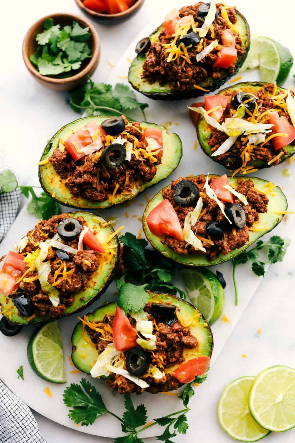 Taco stuffed avocadosย are a healthy twist on tacos and you will love the textures of this flavorful version. Ripe avocados stuffed with seasoned beef and loaded with all your favorite taco toppings! Top view of taco stuffed avocados on a white marble slab. The avocados are garnished with tomatoes, olives, cheese, and cilantro.