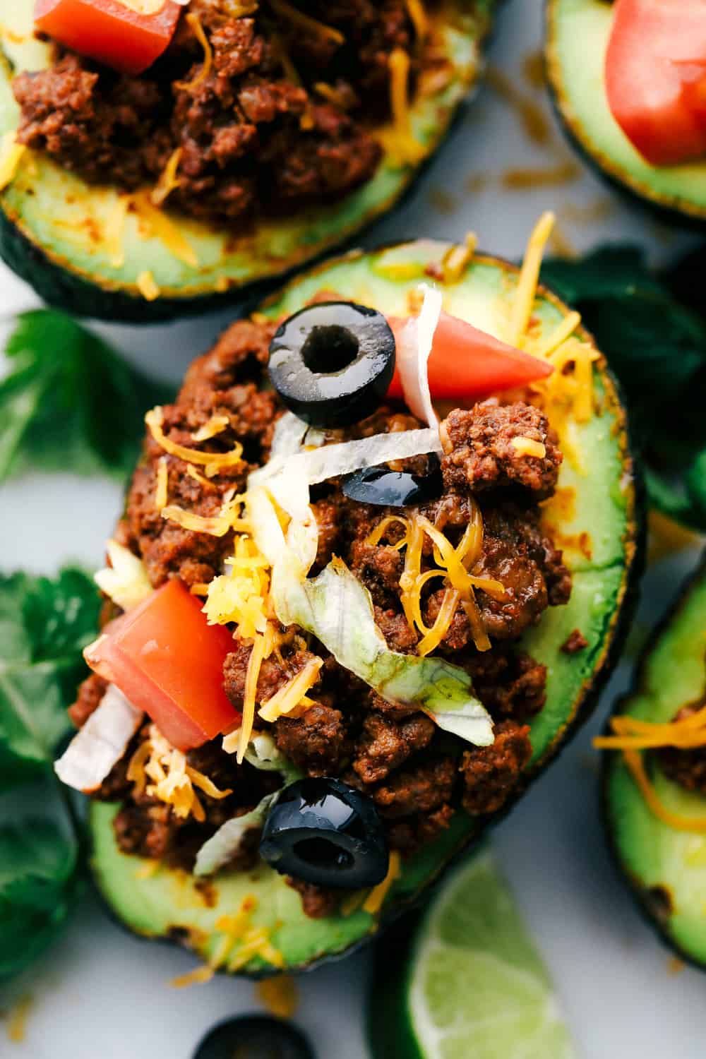 Taco stuffed avocadosย are a healthy twist on tacos and you will love the textures of this flavorful version. Ripe avocados stuffed with seasoned beef and loaded with all your favorite taco toppings! Close over head view of taco stuffed avocados on a white marble slab. The avocados are garnished with tomatoes, olives, cheese, and cilantro.