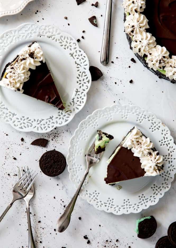 A creamy Mint Oreo Cheesecake served on a crisp Oreo cookie crust and made with no waterbath required! Top it off with a dark chocolate ganache and whipped cream for a decadent mint and chocolate treat! Two slices of Mint Oreo Cheesecake on white plates with metal forks. There are also mint Oreos on the side.