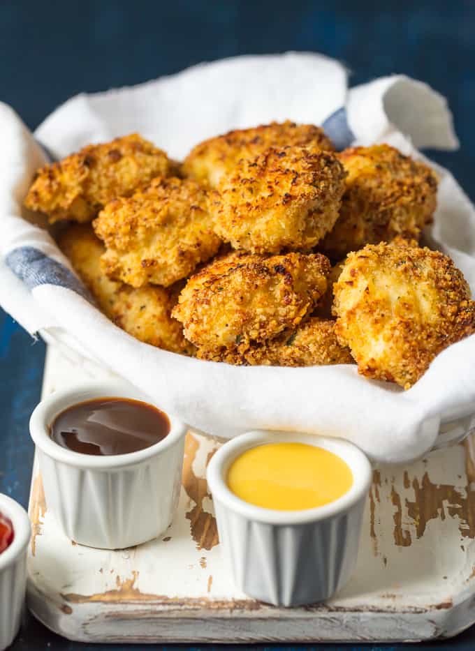 Theseย Baked Parmesan Chicken Nuggets are the perfect easy and healthy meal your entire family will love. Baked Parmesan Chicken Nuggets in a bowl on a cutting board. There are three different types of dipping sauce also.