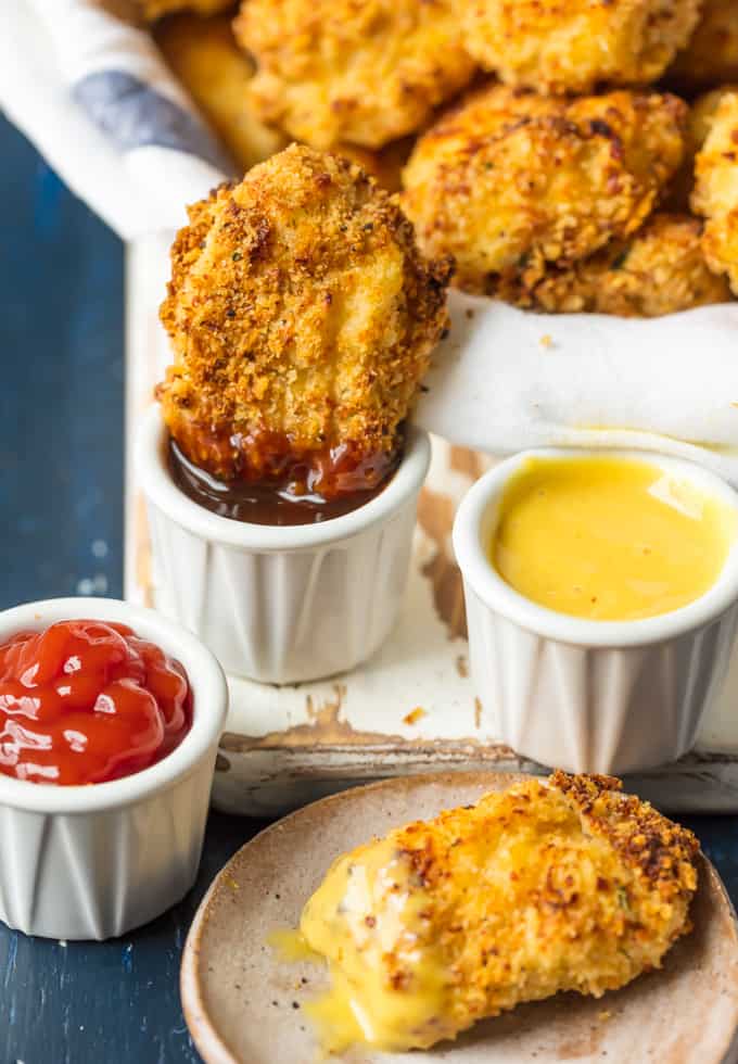 Theseย Baked Parmesan Chicken Nuggets are the perfect easy and healthy meal your entire family will love. A closeup of Baked Parmesan Chicken Nuggets. One being dipped in BBQ sauce and one being dipped in honey mustard. There is also a small dish of ketchup.