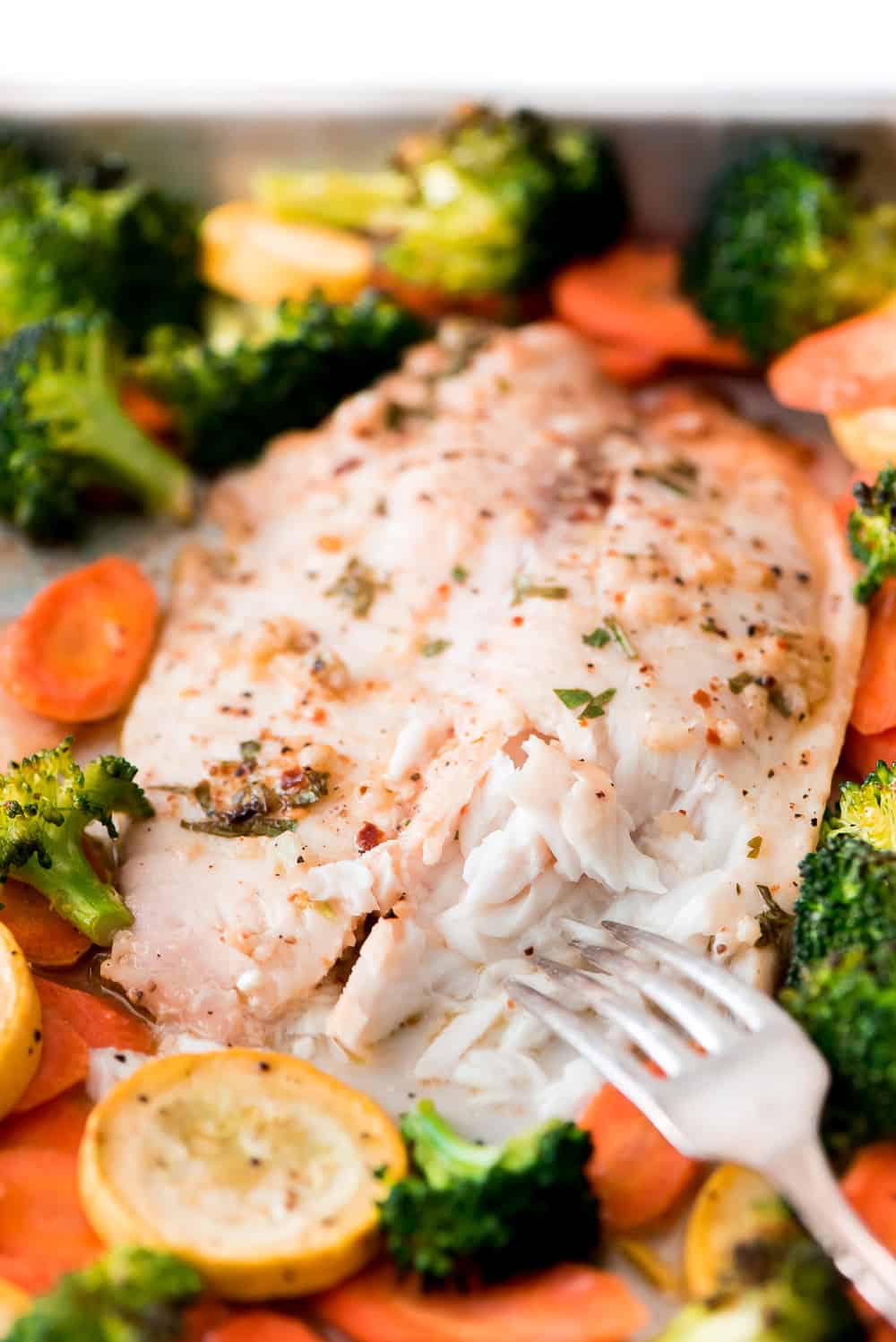 Baked Tilapia and Roasted Veggies is a low calorie and high protein dish that comes together in less than 25 minutes! Light and tasty in every bite!ย A fork pulling a bite of Baked Tilapia.