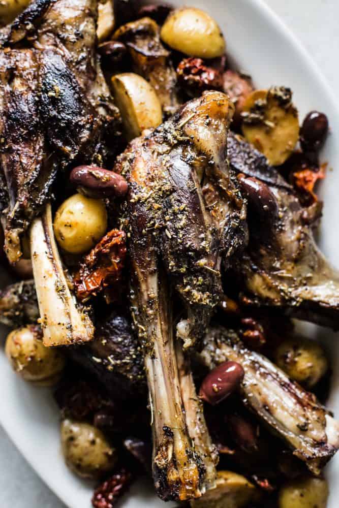 Slow Cooker Greek Lamb Shanks are fall off the bone tender and rubbed in the best greek spices. Potatoes, sun-dried tomatoes, and kalamata olives all cook to perfection in the slow cooker and make such a delicious meal in one!ย Close-up of Slow Cooker Greek Lamb Shanks on a white plate.
