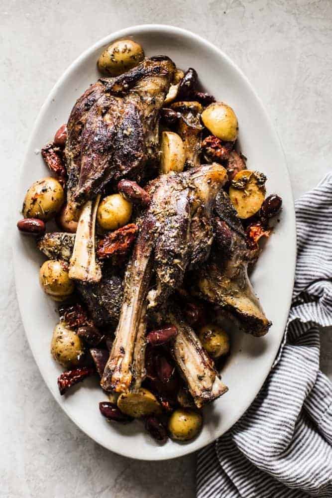 Slow Cooker Greek Lamb Shanks are fall off the bone tender and rubbed in the best greek spices. Potatoes, sun-dried tomatoes, and kalamata olives all cook to perfection in the slow cooker and make such a delicious meal in one!ย Slow Cooker Greek Lamb Shanks on a white platter.