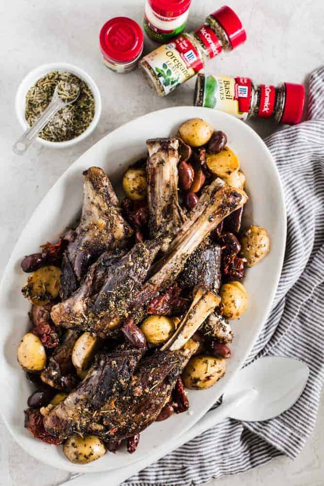 Slow Cooker Greek Lamb Shanks are fall off the bone tender and rubbed in the best greek spices. Potatoes, sun-dried tomatoes, and kalamata olives all cook to perfection in the slow cooker and make such a delicious meal in one!ย Slow Cooker Greek Lamb Shanks on a white platter with McCormick seasonings.