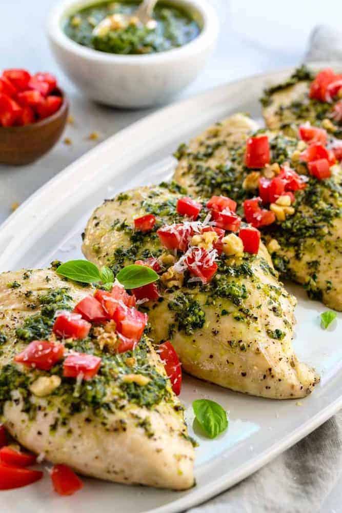 Pesto Chicken is a healthy baked dish that is loaded with flavor. The pesto sauce is made with fresh basil, spinach, garlic, walnuts and Parmesan cheese that gets added right on top of the tender chicken to make each bite irresistible! Family-style pesto chicken dinner on a white platter.
