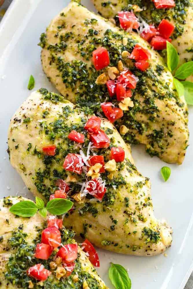 Pesto Chicken is a healthy baked dish that is loaded with flavor. The pesto sauce is made with fresh basil, spinach, garlic, walnuts and Parmesan cheese that gets added right on top of the tender chicken to make each bite irresistible! Pesto chicken breasts on a white serving plate