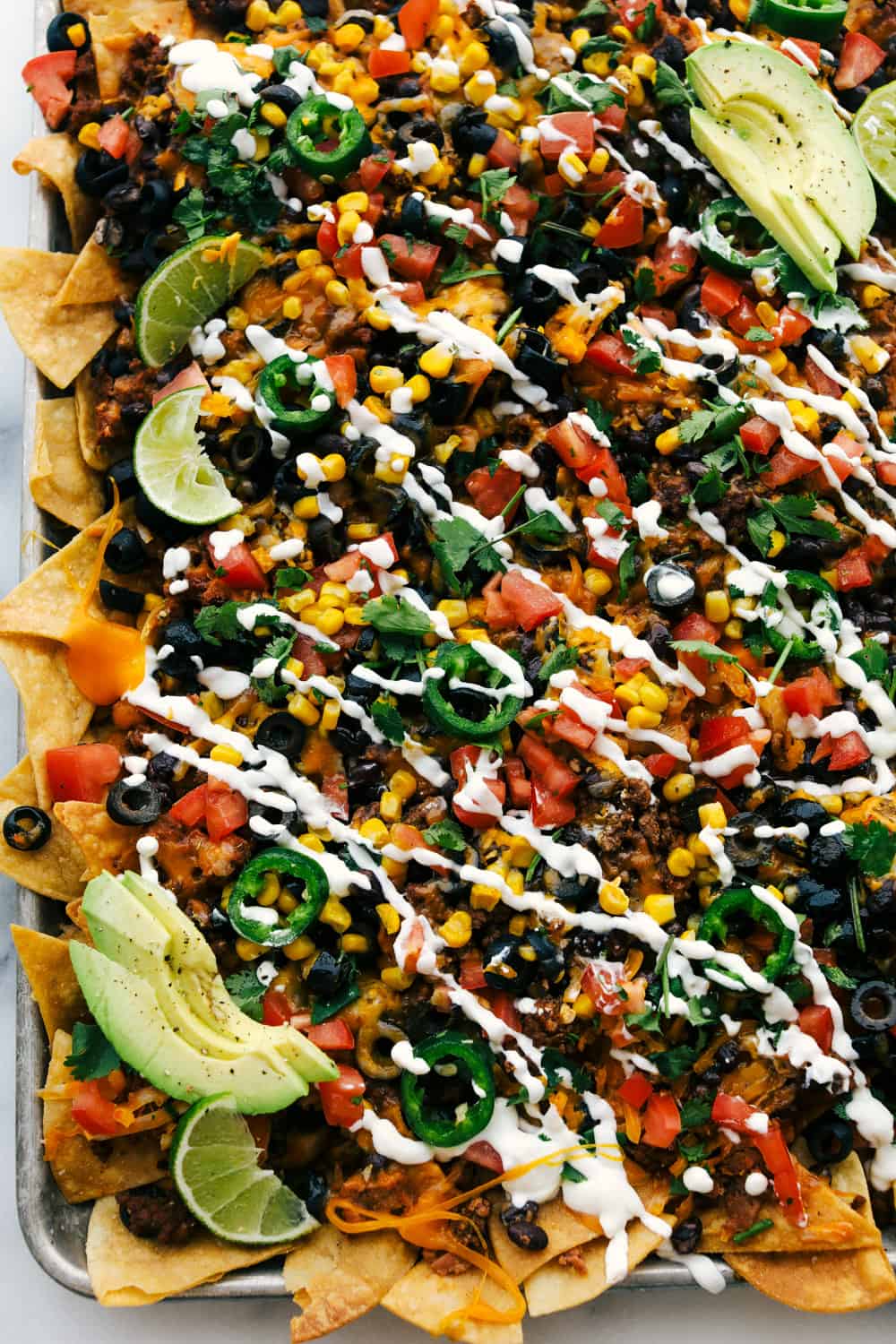 Oven Baked Sheet Pan Nachos Recipe are filled with a layer of chips, ground beef, refried beans, black olives, chopped tomatoes, corn, cheese sprinkled over top then baked to perfection! The possibilities are endless! Oven baked sheet pan nachos with sour cream and avocado over top.