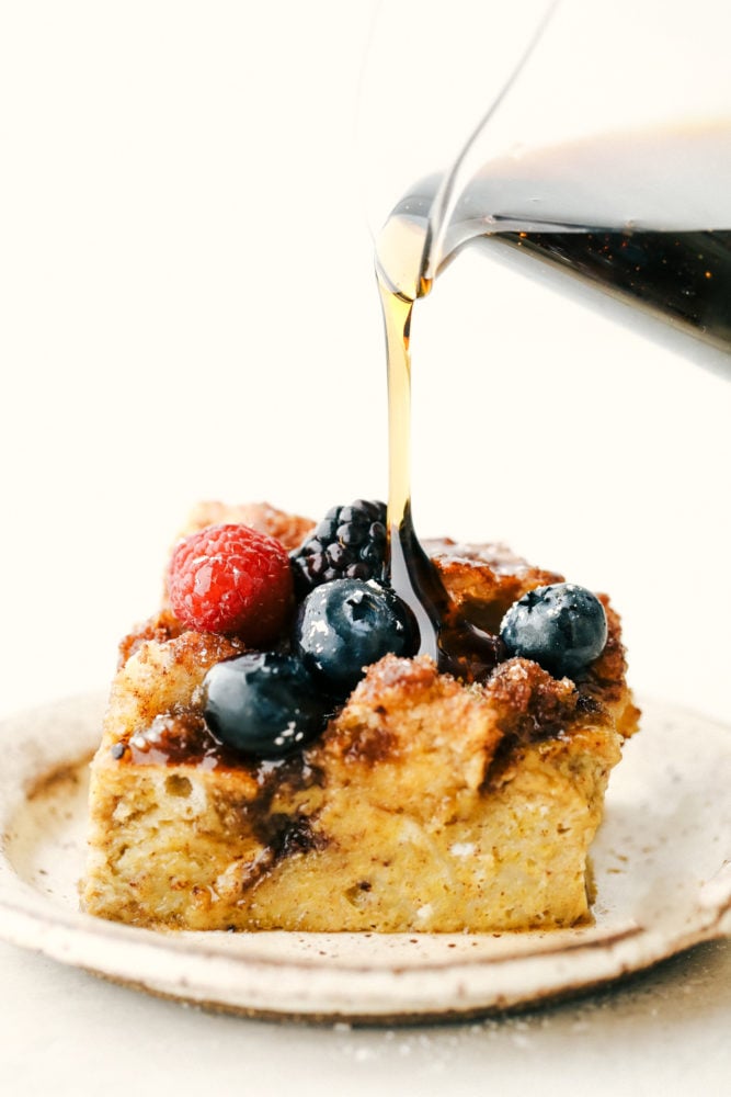 Cinnamon baked French toast is a delicious breakfast casserole full of cinnamon sugar goodness! Soft brioche bread pieces get baked in a sweet creamy custard. French toast has never been easier! Baked French toast slice on a plate with syrup being poured overtop.