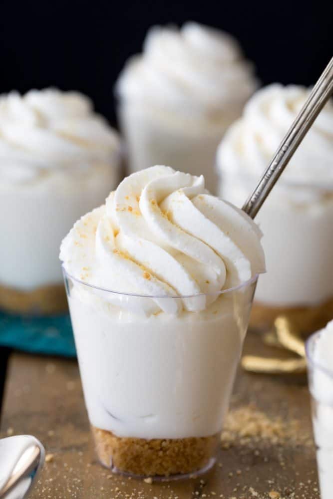 Cheesecake Dessert Cups are served in single-serving-sized 2-oz shot glasses and are made with a simple graham cracker crust and a creamy no-bake cheesecake filling! Finish yours off with your favorite toppings including whipped cream or fruit pie filling! Cheesecake dessert cup topped with homemade whipped cream.
